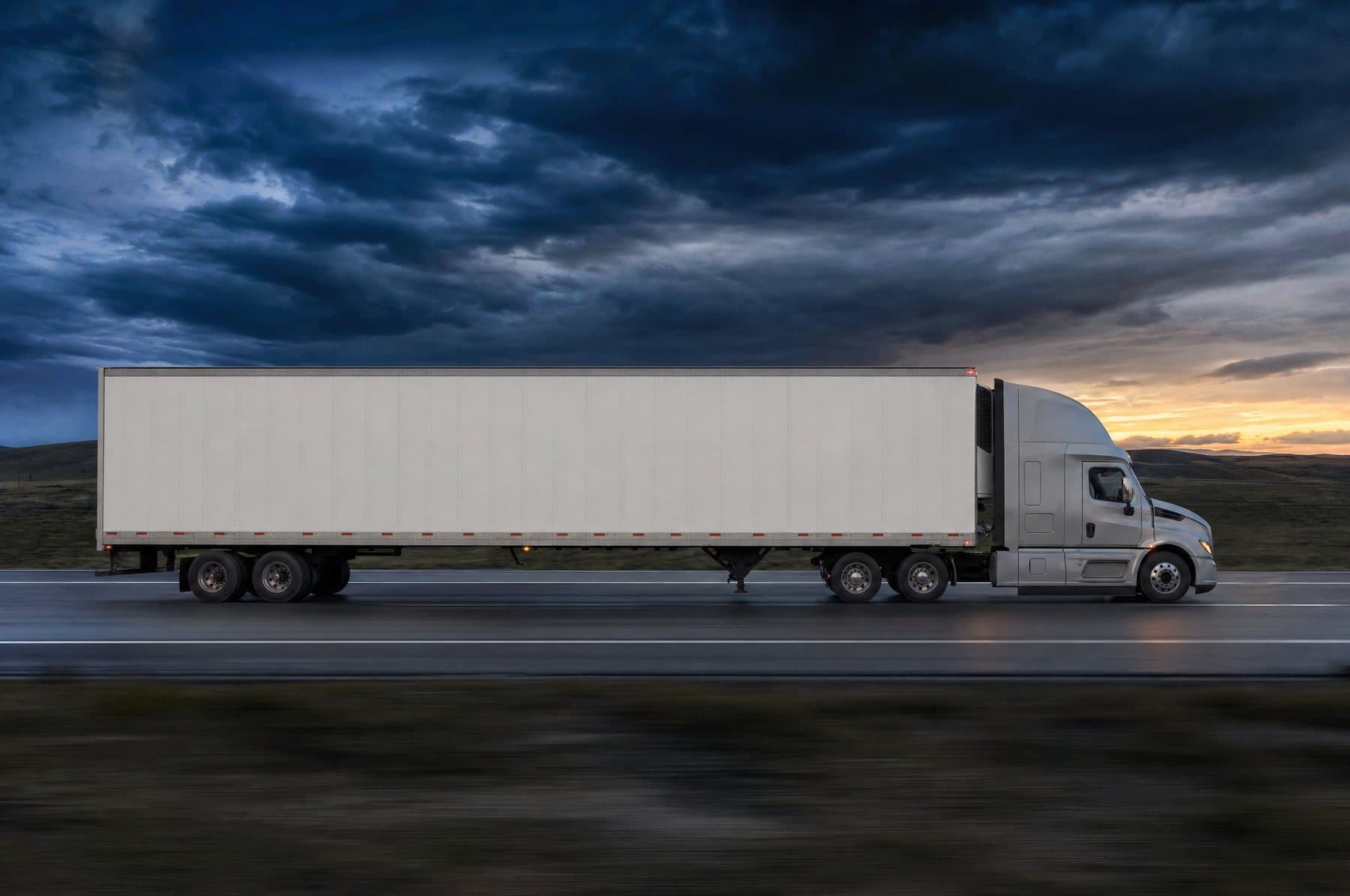 Tractor pulling an unmarked white dry van trailer along an interstate at dusk